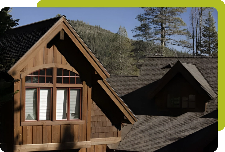 A wooden house with a gabled roof, crafted with the expertise of top roofing companies, sits serenely surrounded by trees and majestic mountains in the background.